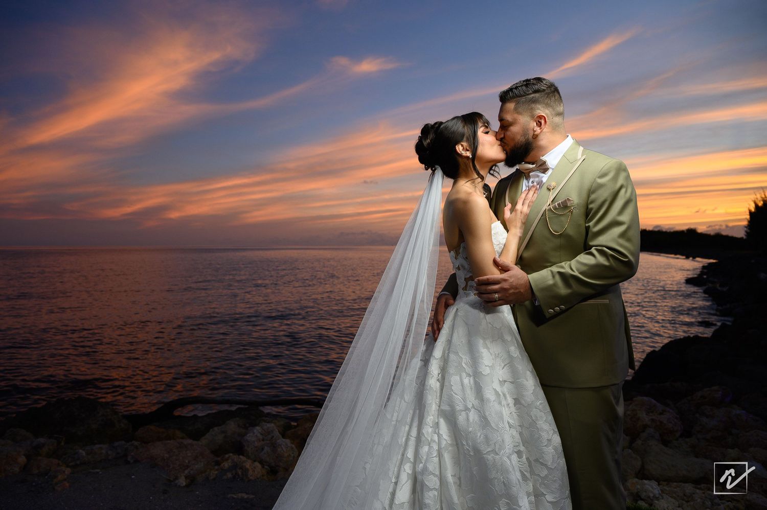 Bride in a white gown and long veil kisses her groom in a green suit on a rocky shore at sunset with a colorful sky over the water.