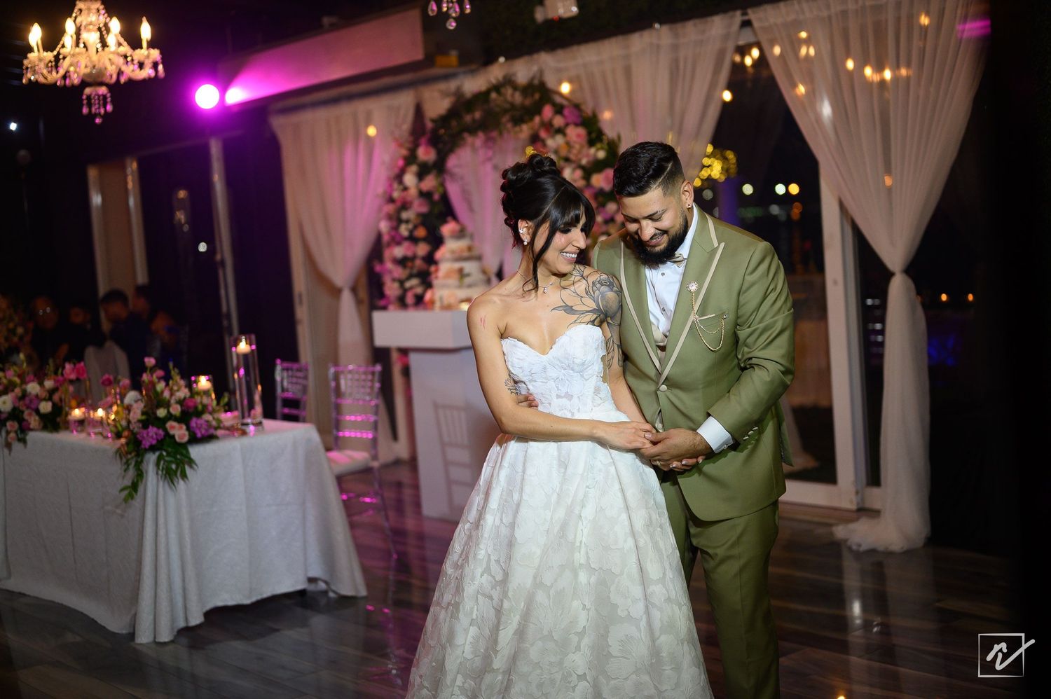 Bride in a white strapless lace gown and groom in an olive green suit share a first dance at a romantic wedding reception with floral decor.
