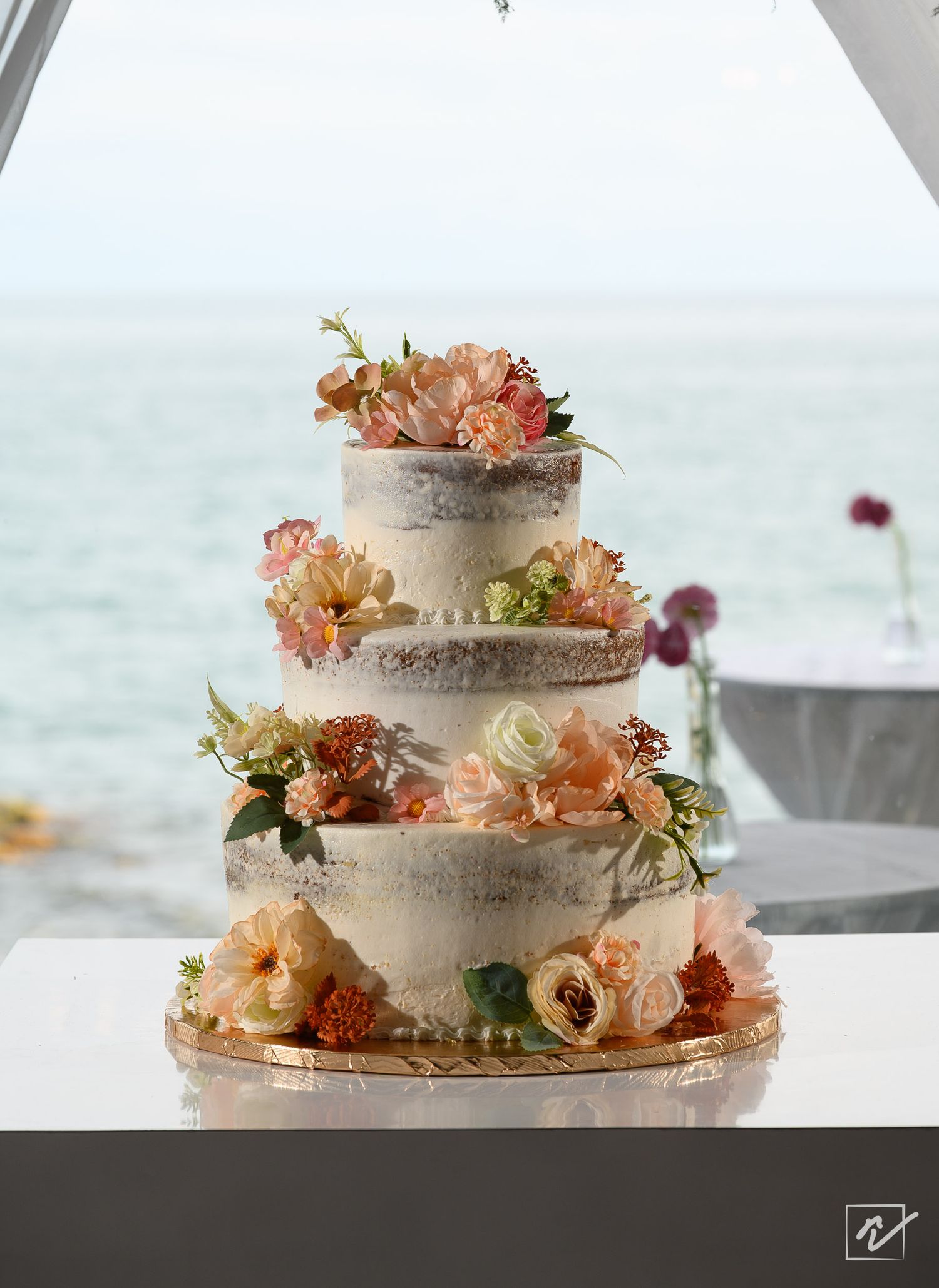 Three-tier naked wedding cake decorated with peach, coral and cream fresh flowers on a gold board, set against a calm ocean backdrop.