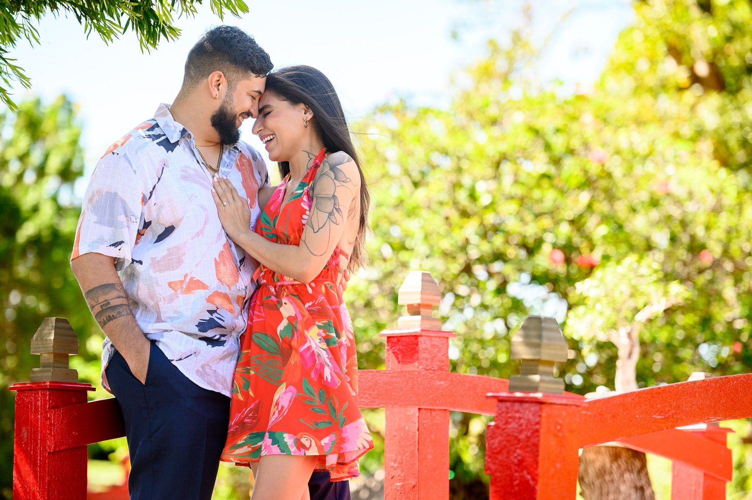 A couple shares a romantic moment on a red bridge surrounded by lush green trees in a vibrant outdoor setting.