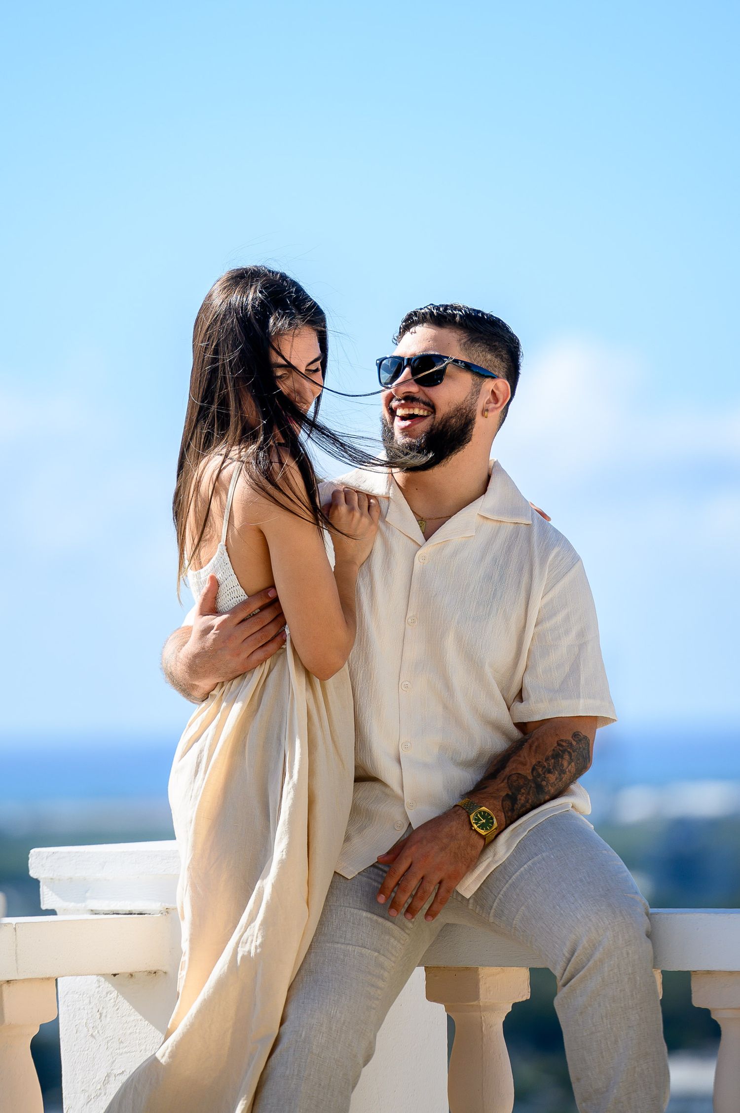 Happy couple in matching cream outfits embracing on sunny outdoor terrace with ocean view.