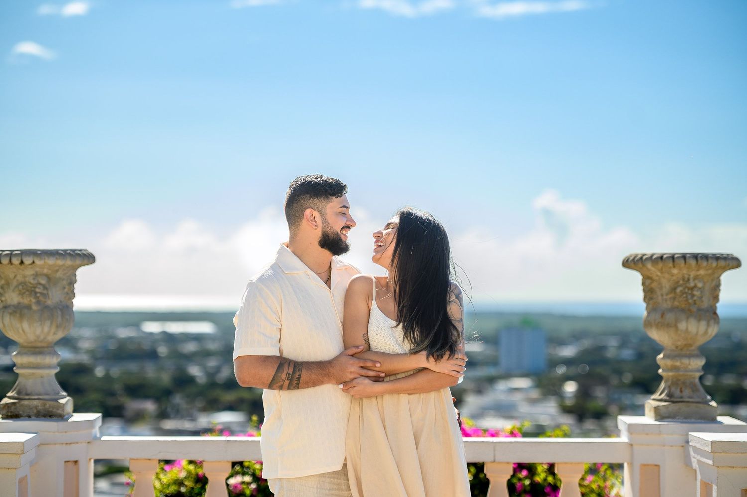 Couple sharing a tender moment on a white balcony with decorative urns and panoramic city views behind them.