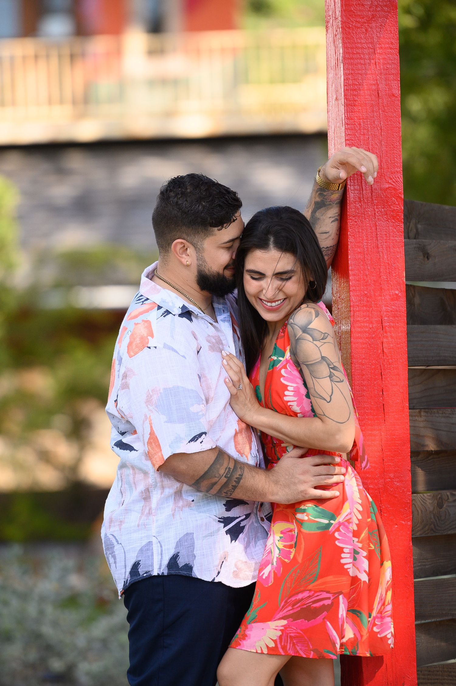 A smiling couple embraces near a red wooden post outdoors, both wearing floral outfits and tattoos on a sunny day.