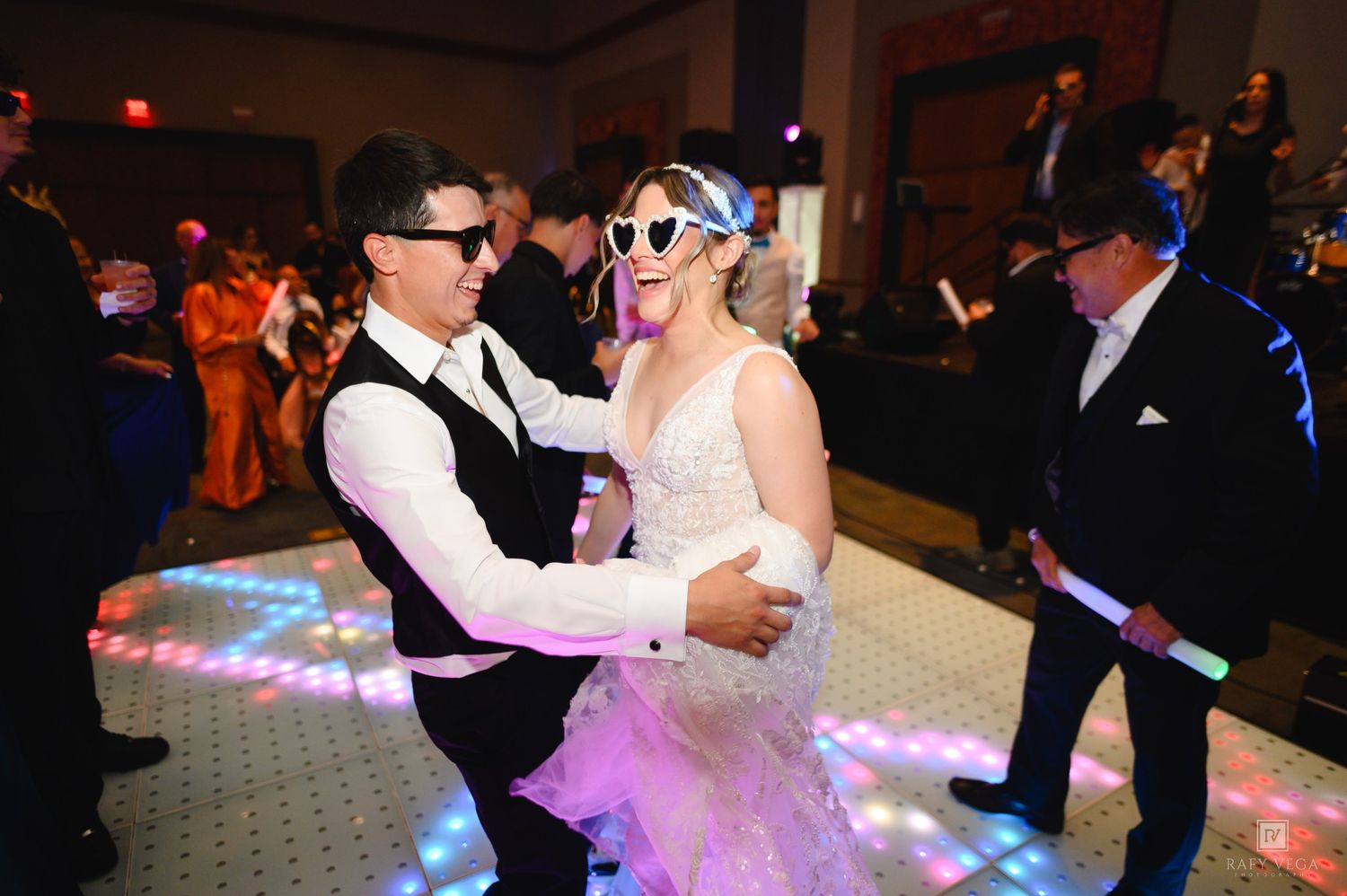 Bride and groom dancing at wedding reception on LED dance floor wearing fun sunglasses with guests in background.