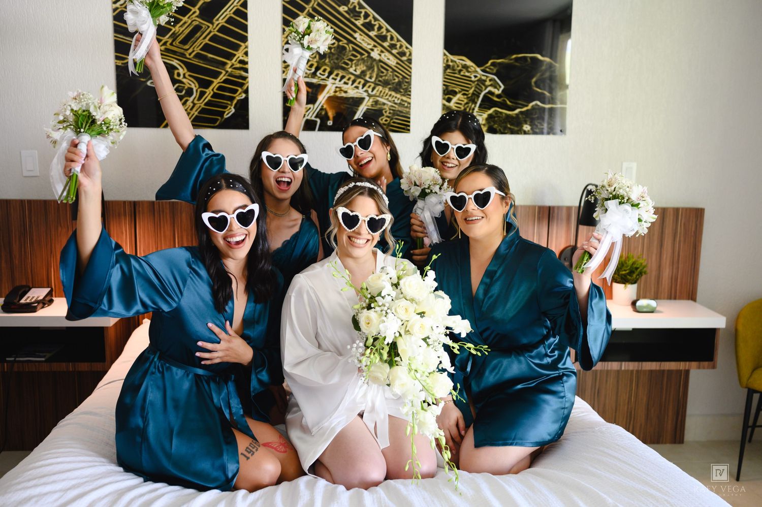 Bride and bridesmaids in teal robes and heart sunglasses celebrate on a hotel bed with bouquets.