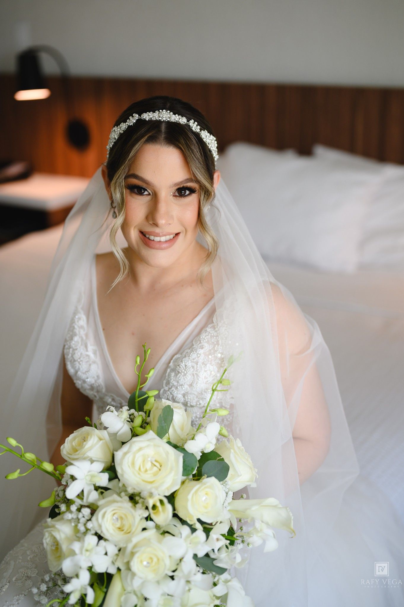 Smiling bride in white lace gown and veil holds elegant white rose bouquet while seated on a bed.
