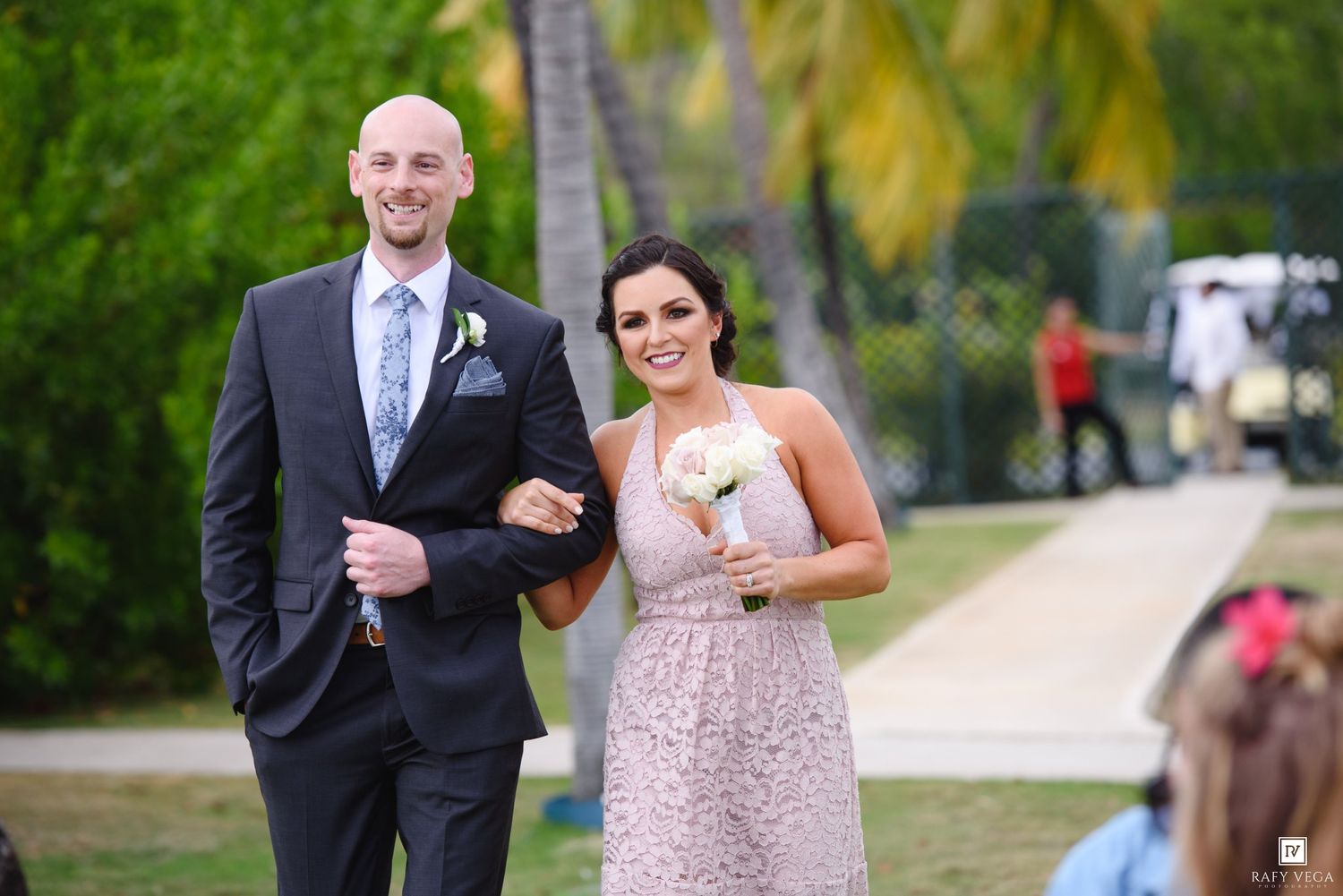 Smiling groomsman in navy suit escorts bridesmaid in pink lace dress down outdoor wedding ceremony aisle.