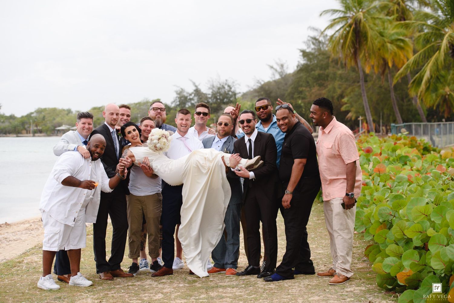 Groom carried by groomsmen in suits on tropical beach during wedding celebration with lush greenery backdrop.
