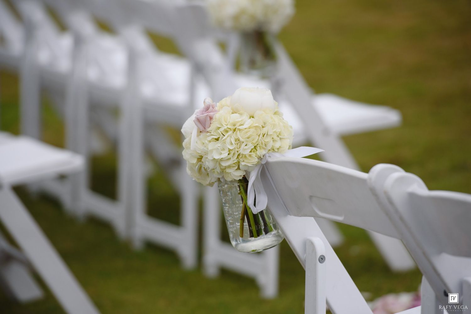 White hydrangea bouquet in mason jar hanging on white folding chair at outdoor wedding ceremony aisle.