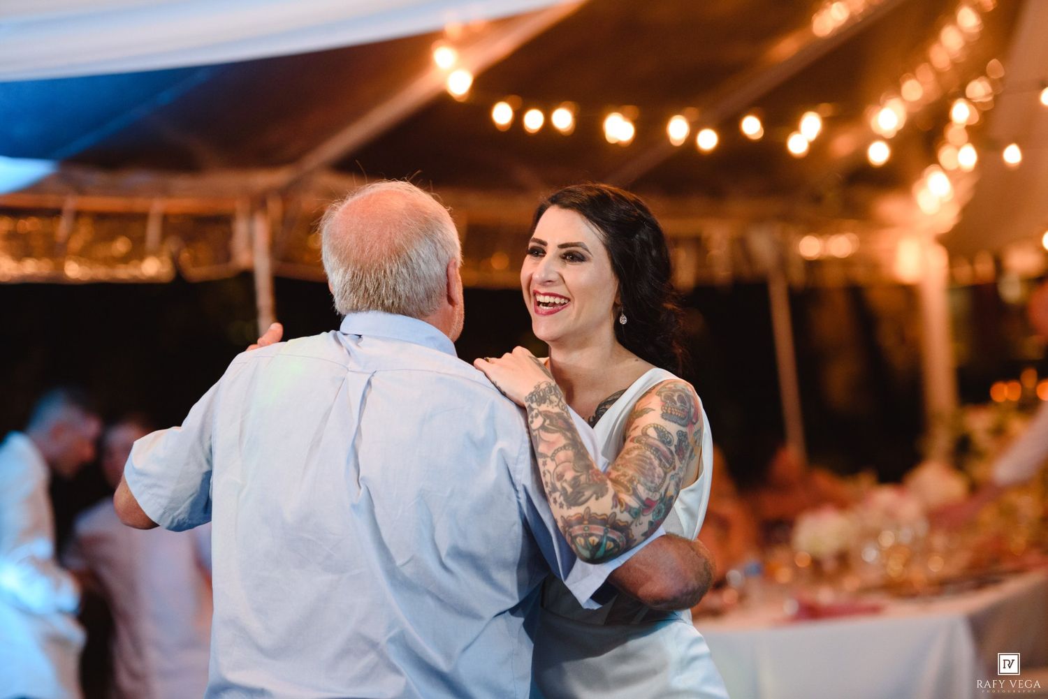 Bride with tattoos laughing while sharing a father-daughter dance under string lights at an outdoor wedding reception.