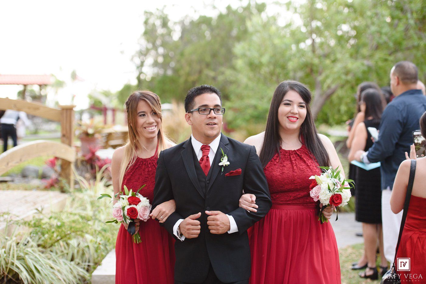 A groom in a black suit escorts two bridesmaids in red dresses down the wedding aisle outdoors.