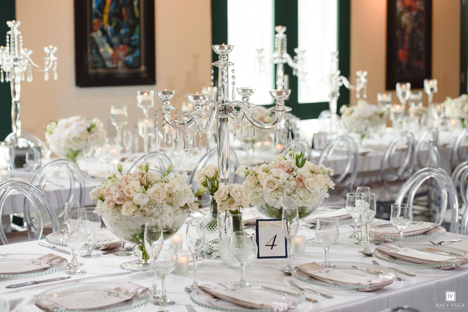 Elegant wedding reception table with crystal candelabras, white floral centerpieces, and ghost chairs in a bright venue.