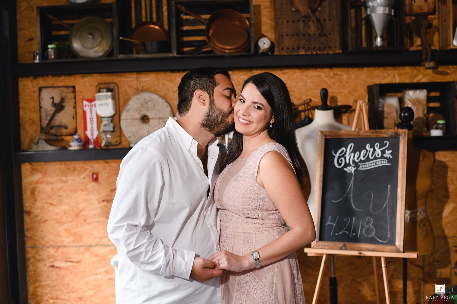Couple sharing a romantic kiss near a chalkboard Cheers sign in a rustic vintage-styled engagement photo.