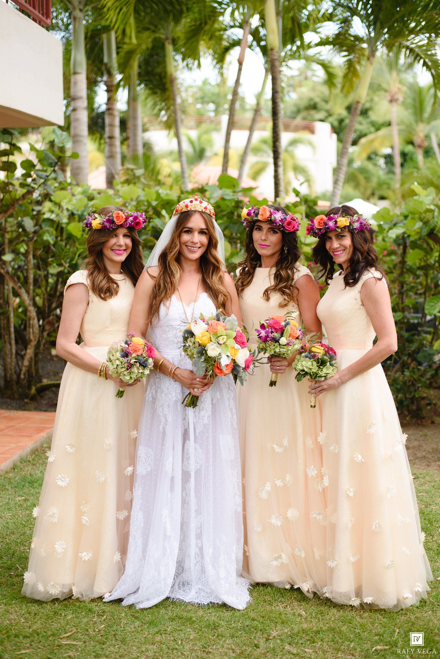 Bride in white lace gown with three bridesmaids in champagne dresses and floral crowns holding colorful bouquets outdoors.