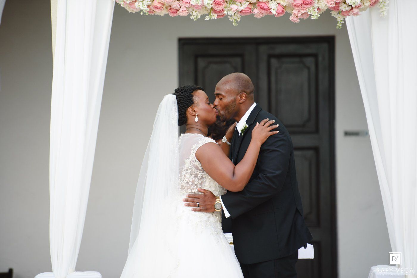 A bride and groom share a romantic first kiss under a floral arch at their outdoor wedding ceremony.
