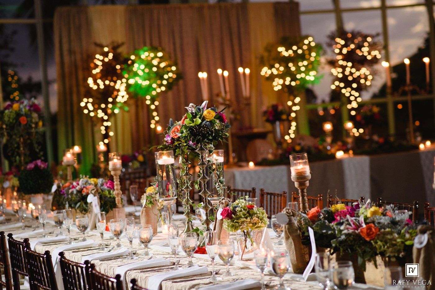 Elegant wedding reception with colorful floral centerpieces, candles, and fairy light trees decorating long dining tables.