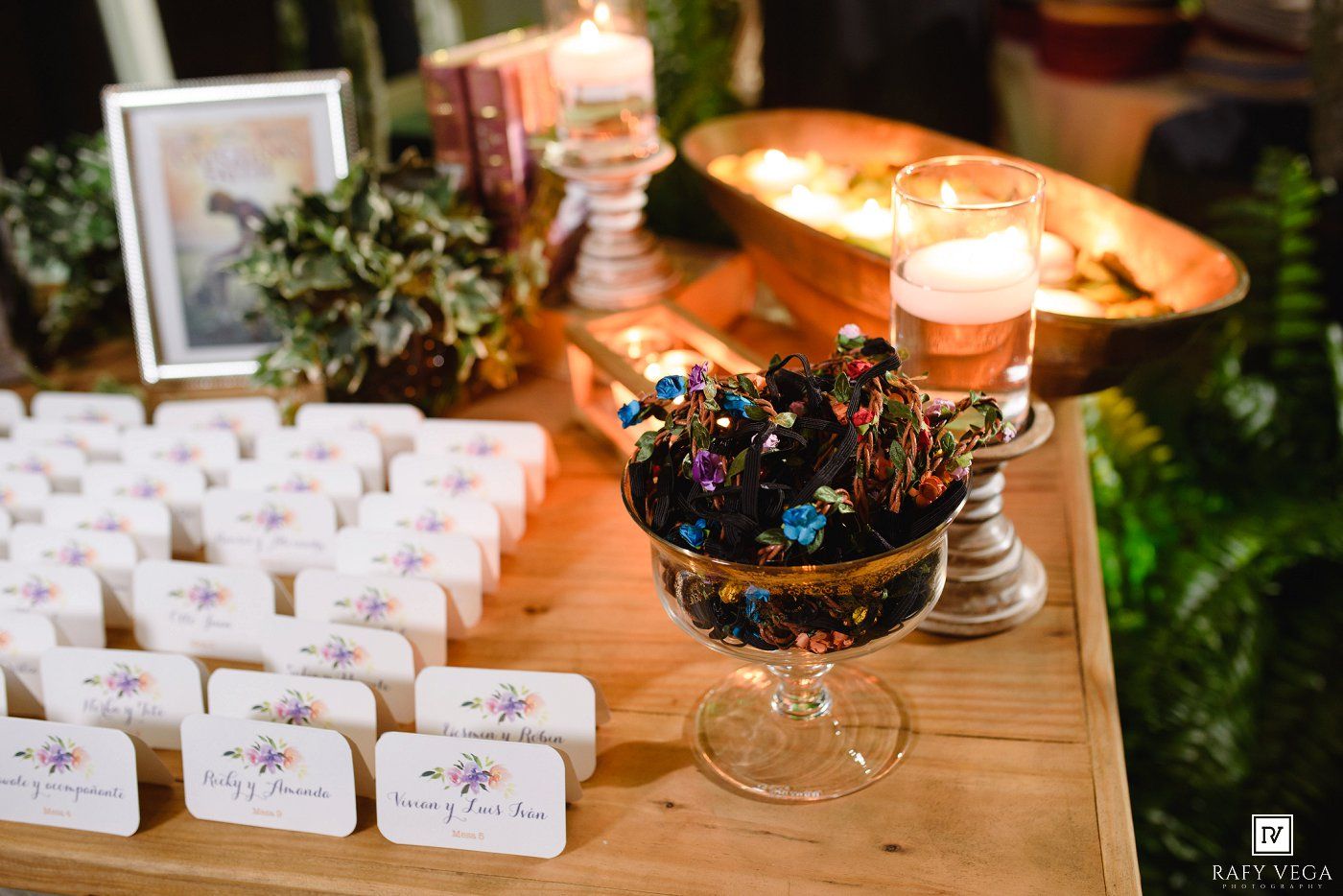 Elegant wedding escort card table with floral place cards, colorful candy bowl, candles, and greenery decor.