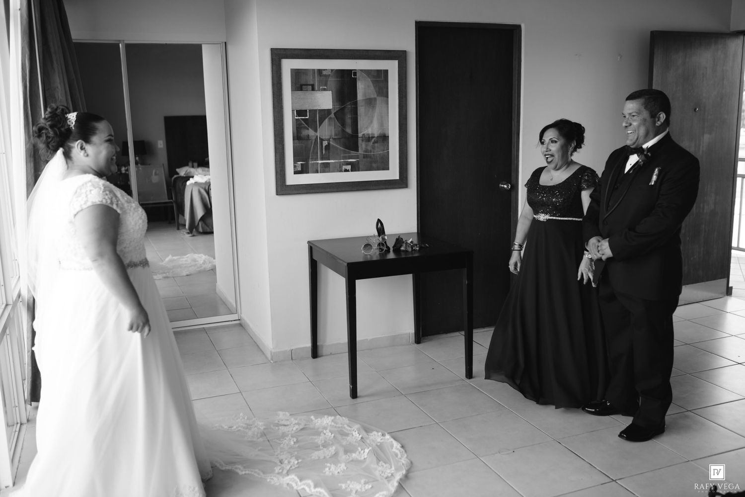 Black and white wedding photo of a bride in a white dress sharing a moment with her parents indoors.