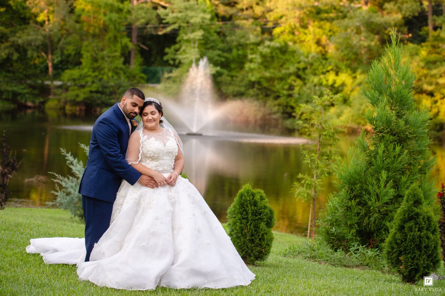 Bride and groom pose elegantly by a serene pond with a fountain surrounded by fall foliage.