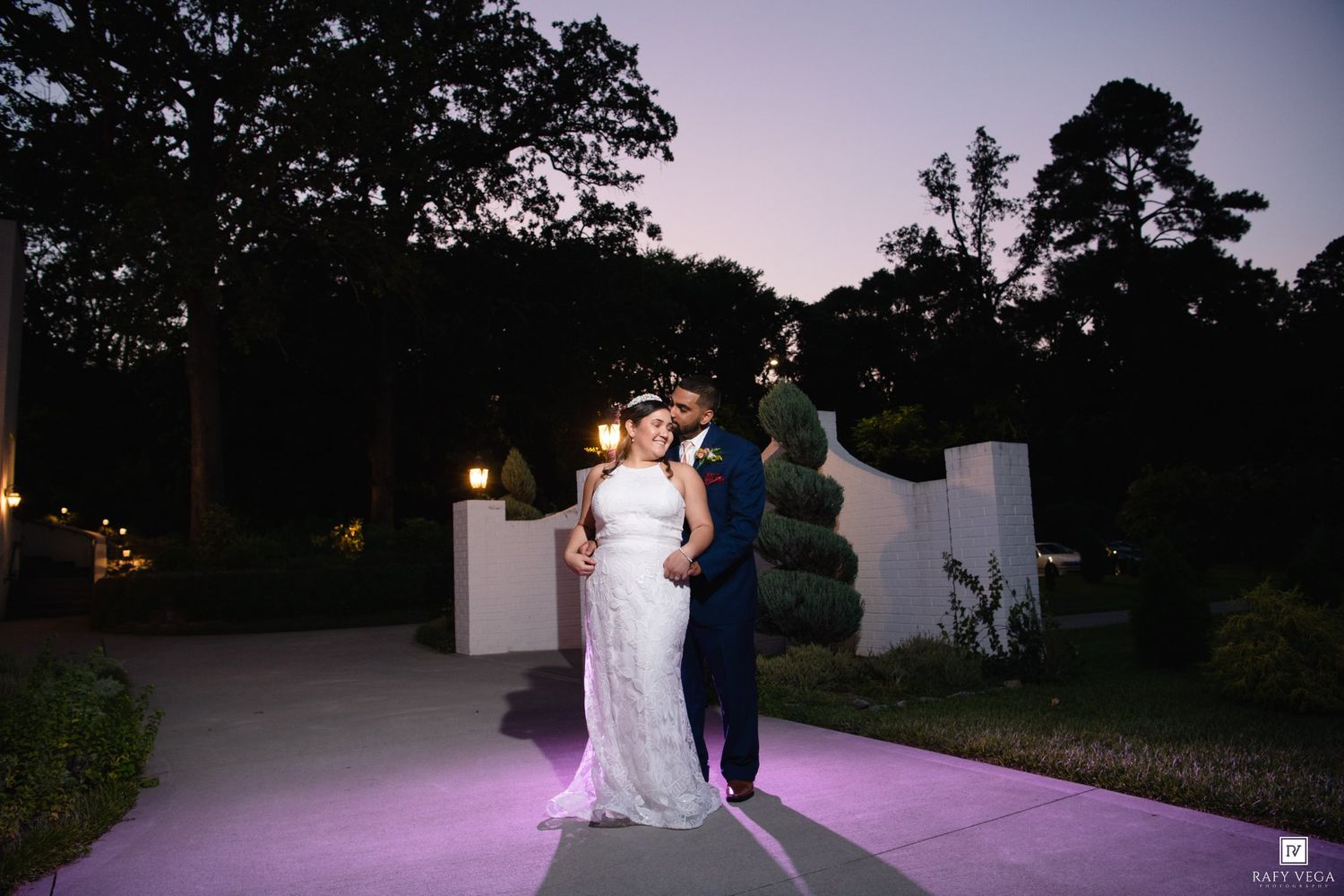 Bride and groom share a romantic kiss at dusk in an elegant garden venue with purple ambient lighting.