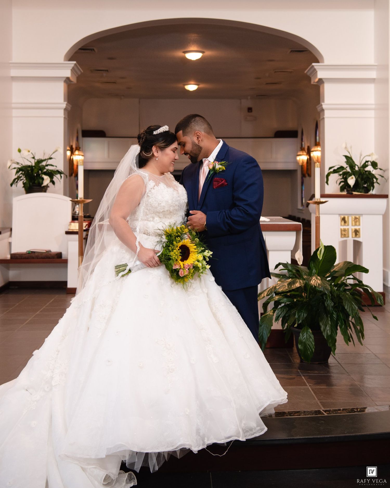 Bride in white ball gown with sunflower bouquet and groom in navy suit share romantic moment at church altar.