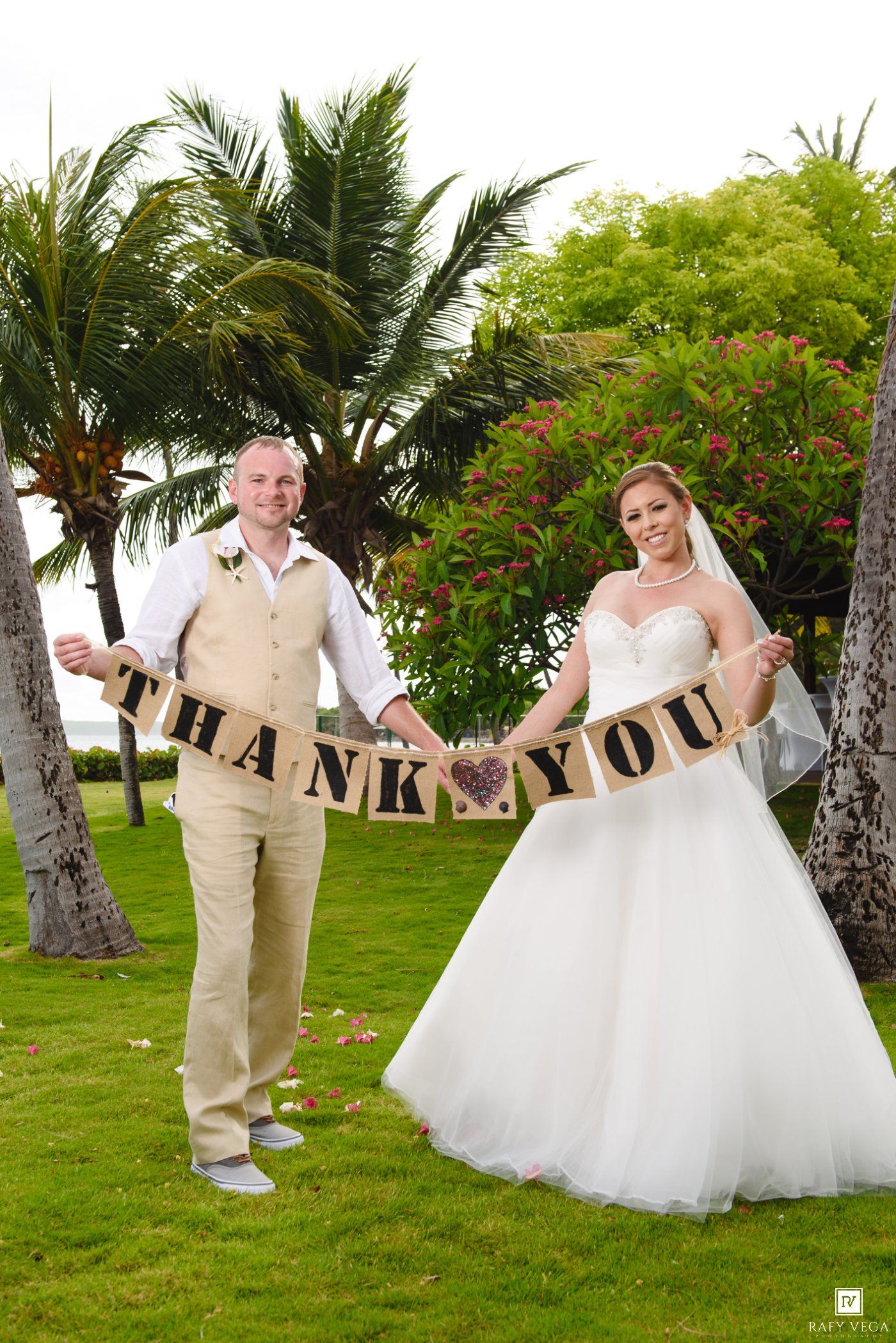 Bride and groom holding a burlap Thank You banner on tropical lawn with palm trees.