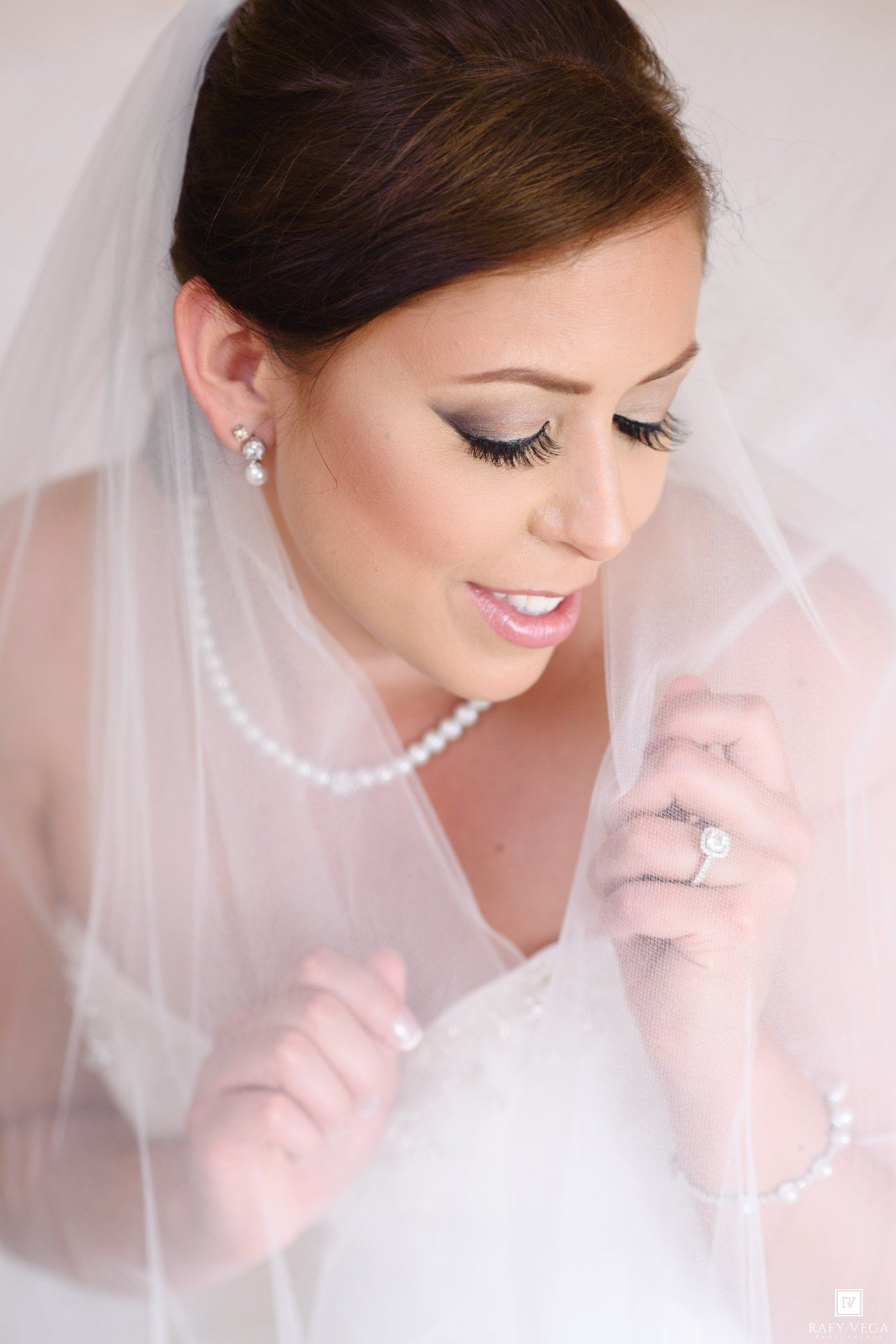 Bride in white veil and pearl necklace with diamond ring, elegant bridal makeup and lashes, looking down softly.