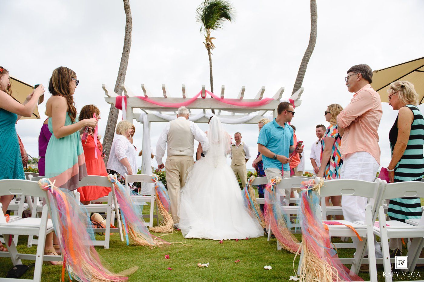 Outdoor beach wedding ceremony with guests seated in white chairs along a floral aisle under a pink decorated arch.