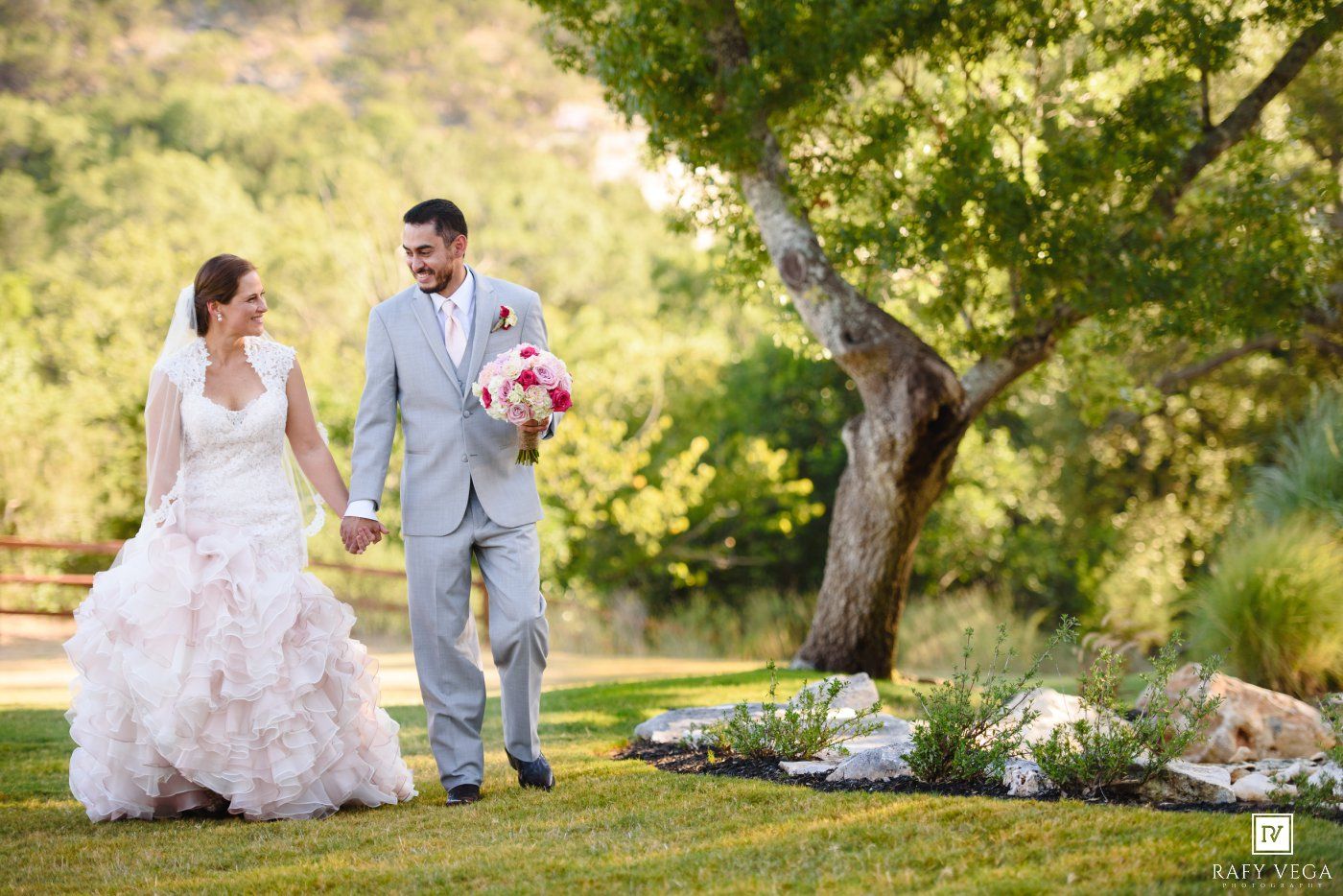 Bride and groom walk hand-in-hand on lush green lawn, bride in ruffled white gown holding pink bouquet.