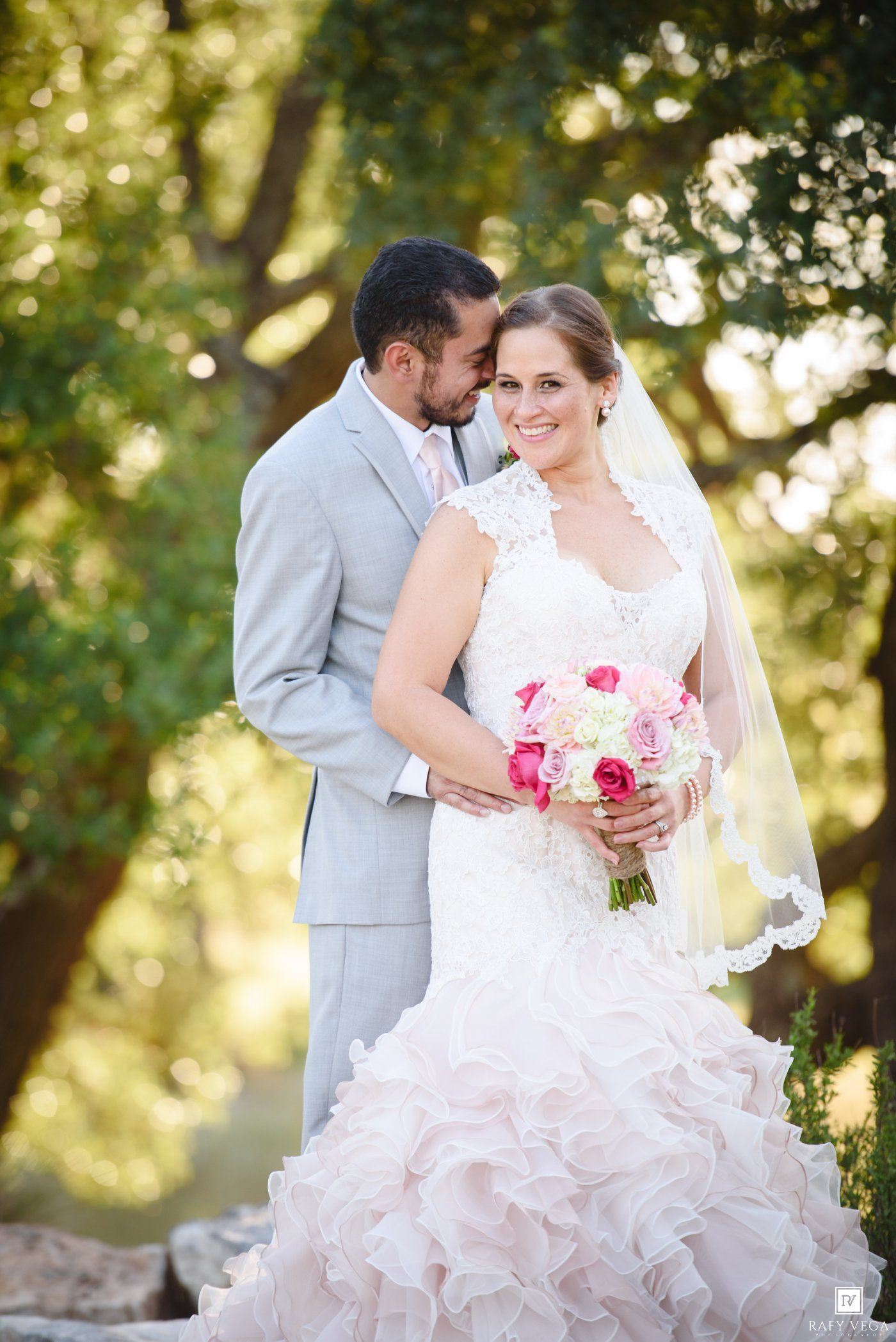 Bride in ruffled white gown holding pink bouquet embraced by groom in gray suit outdoors.