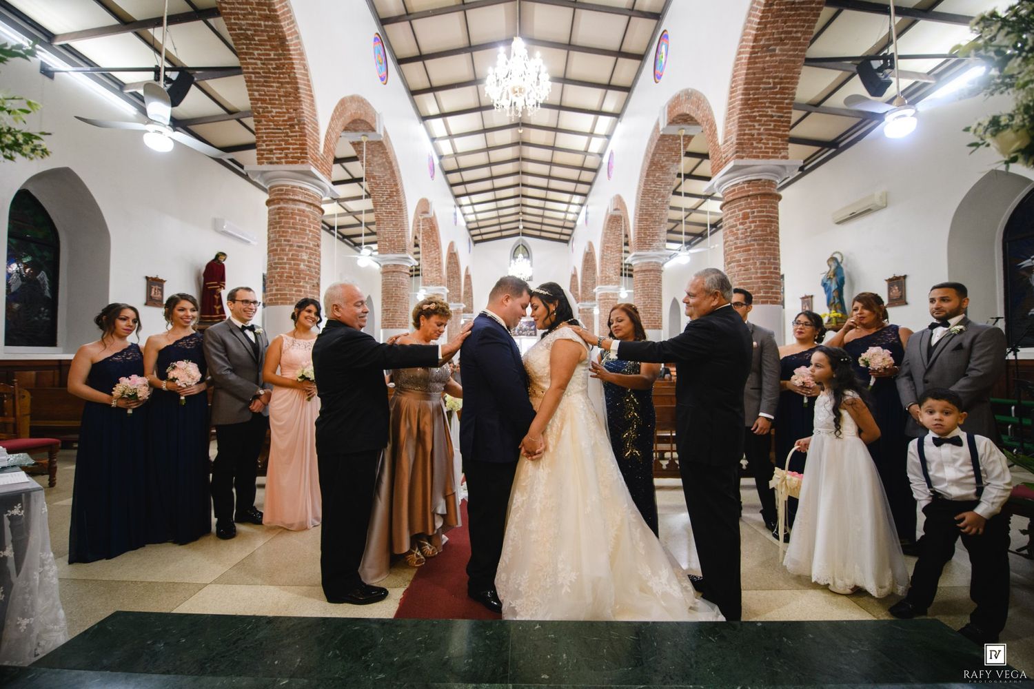 Bride and groom exchange vows at elegant church wedding ceremony surrounded by bridesmaids, groomsmen, and flower girl.