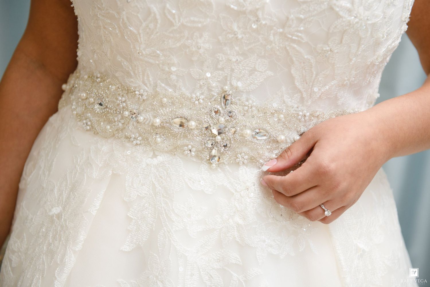 Close-up of a bride wearing a lace wedding gown with a crystal beaded belt, showing her engagement ring.