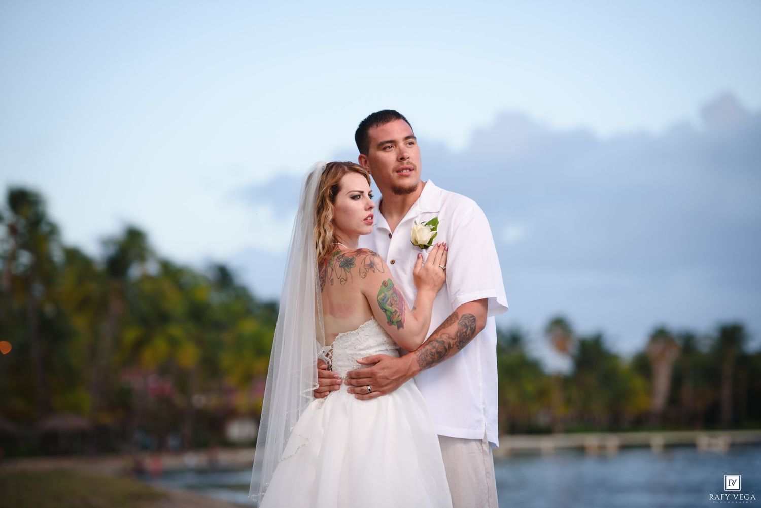 Couple in wedding attire embracing on tropical beach at dusk, bride in white gown with veil, groom in white shirt.