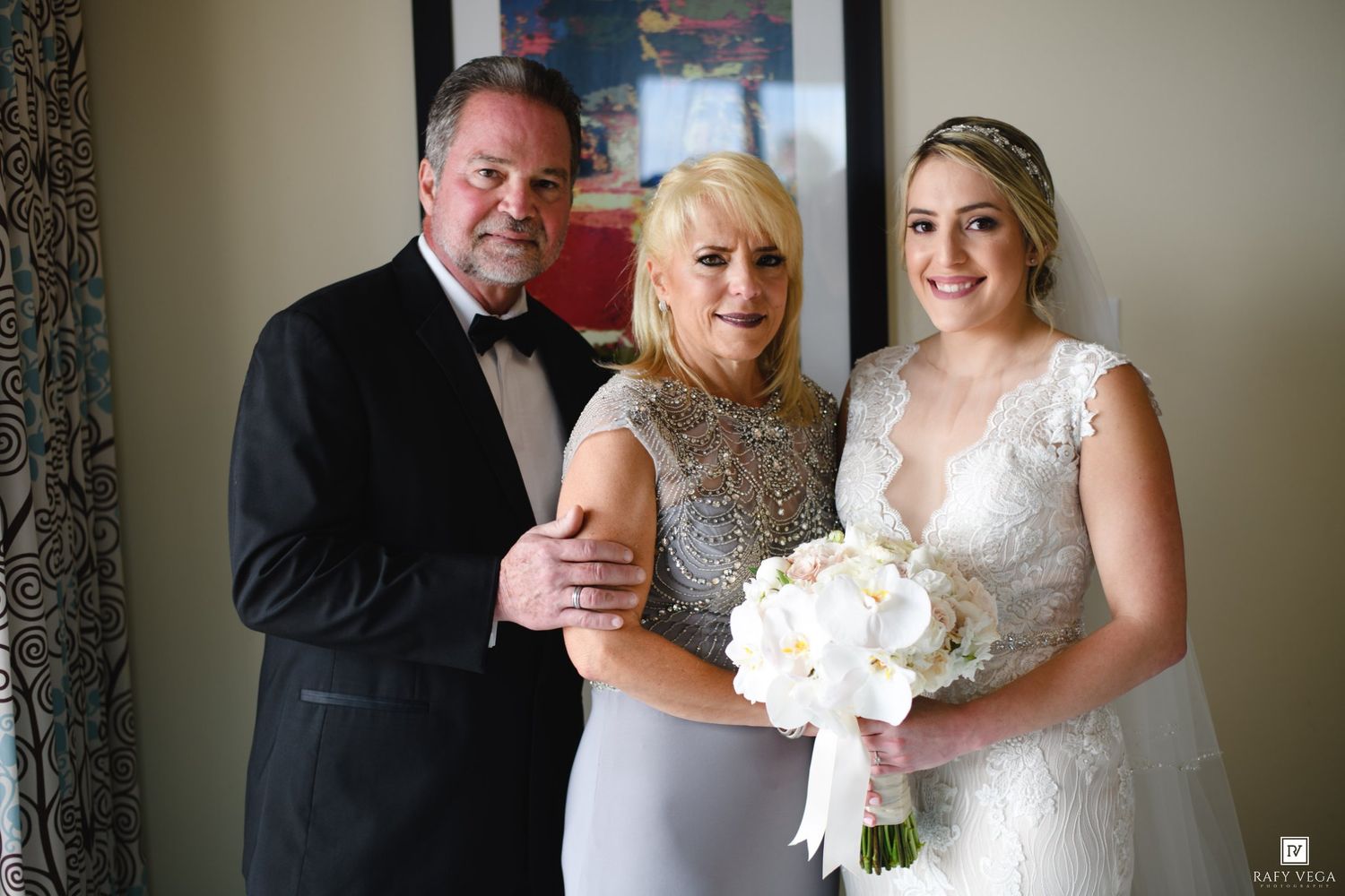 Bride in white lace gown holds bouquet posing with parents in formal attire at wedding.