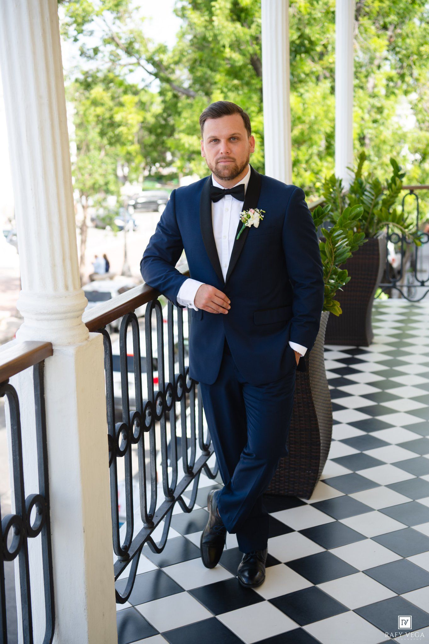 Groom in navy blue tuxedo with bow tie and boutonniere poses on checkered floor balcony.