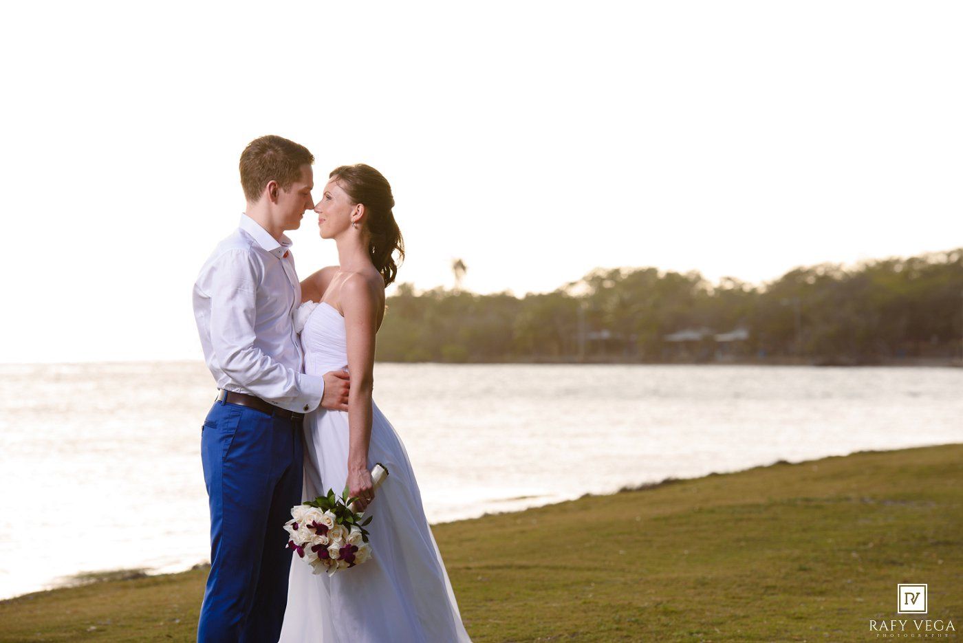 Bride and groom share a romantic moment by the waterfront at sunset, bride holding a bouquet in a white gown.