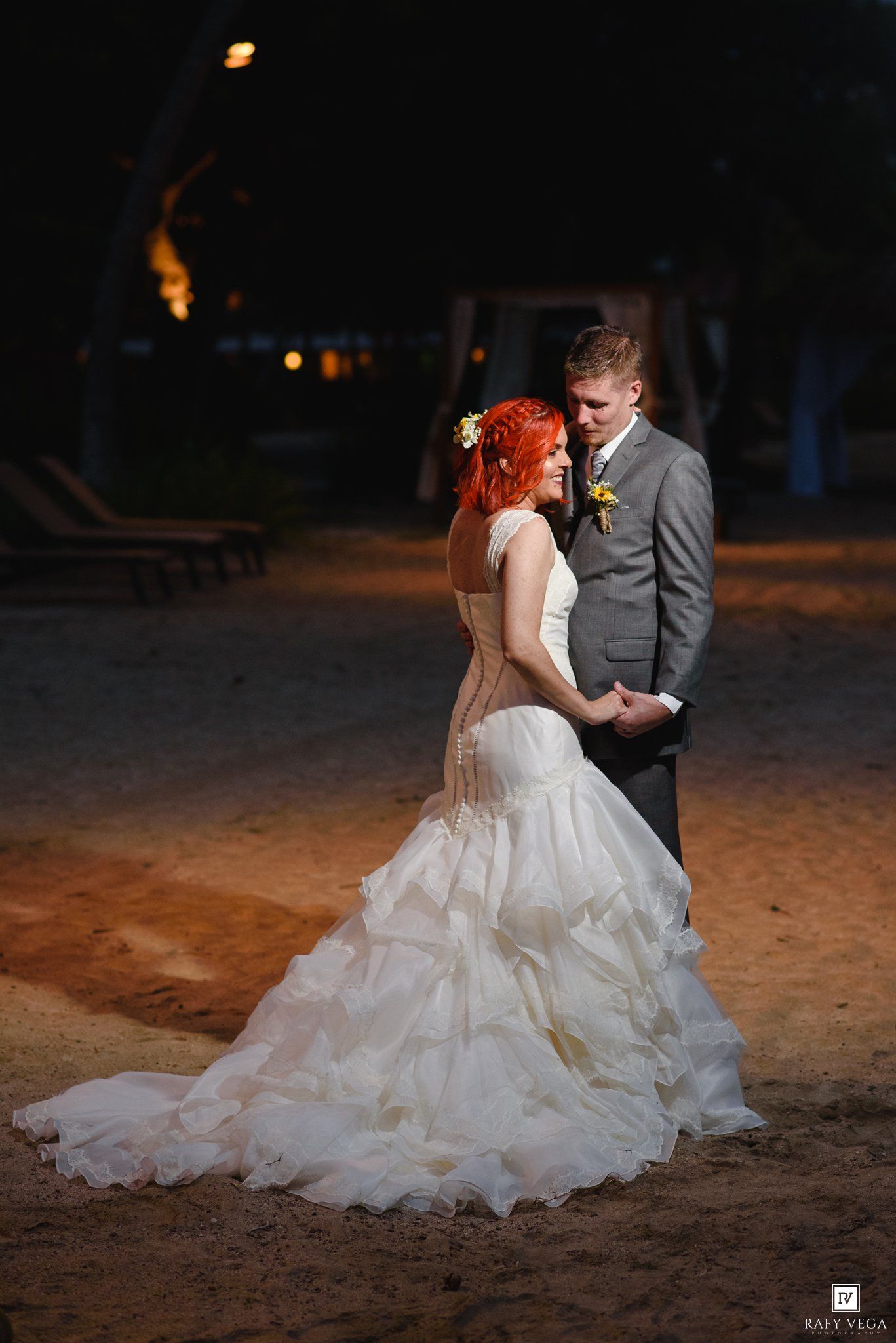 Bride in white gown and groom in gray suit share a romantic moment on sandy beach at night.