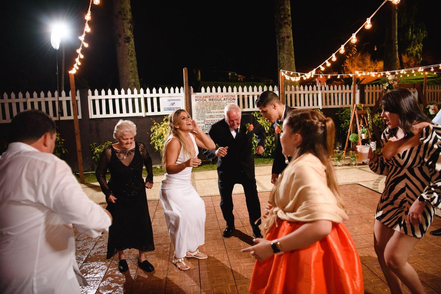 Guests dancing outdoors at a night wedding reception with string lights and white fence backdrop.