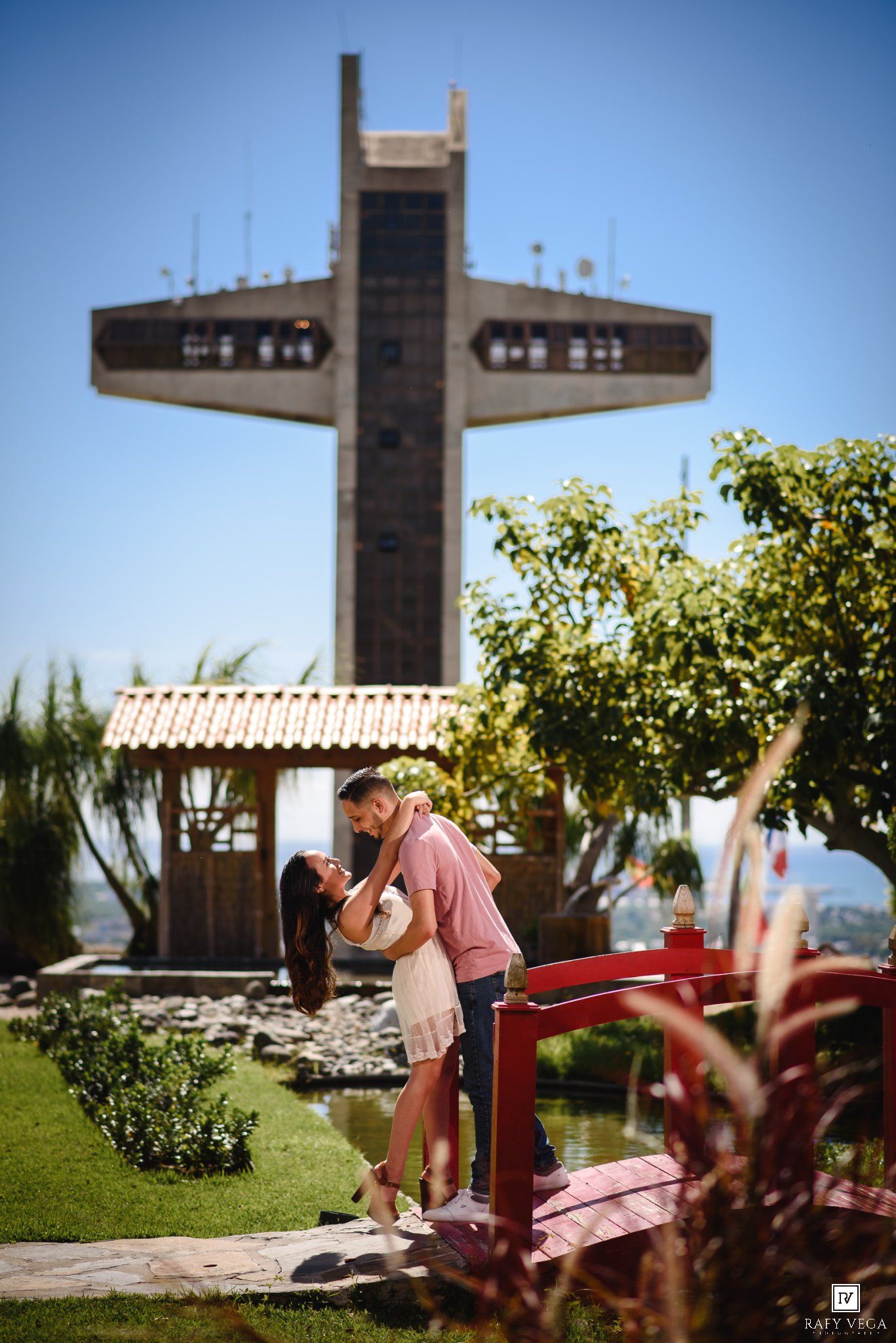 Couple sharing a romantic kiss on a red bridge with a large cross monument and lush garden in the background.