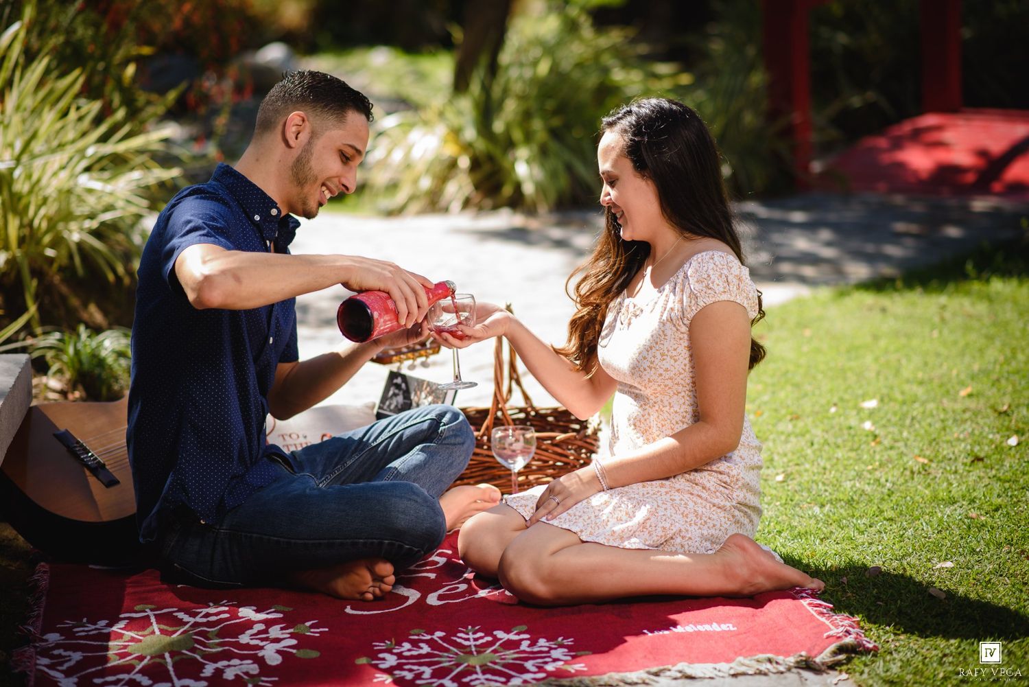 Couple enjoying a romantic outdoor picnic on a red blanket, sharing wine in a lush garden setting.