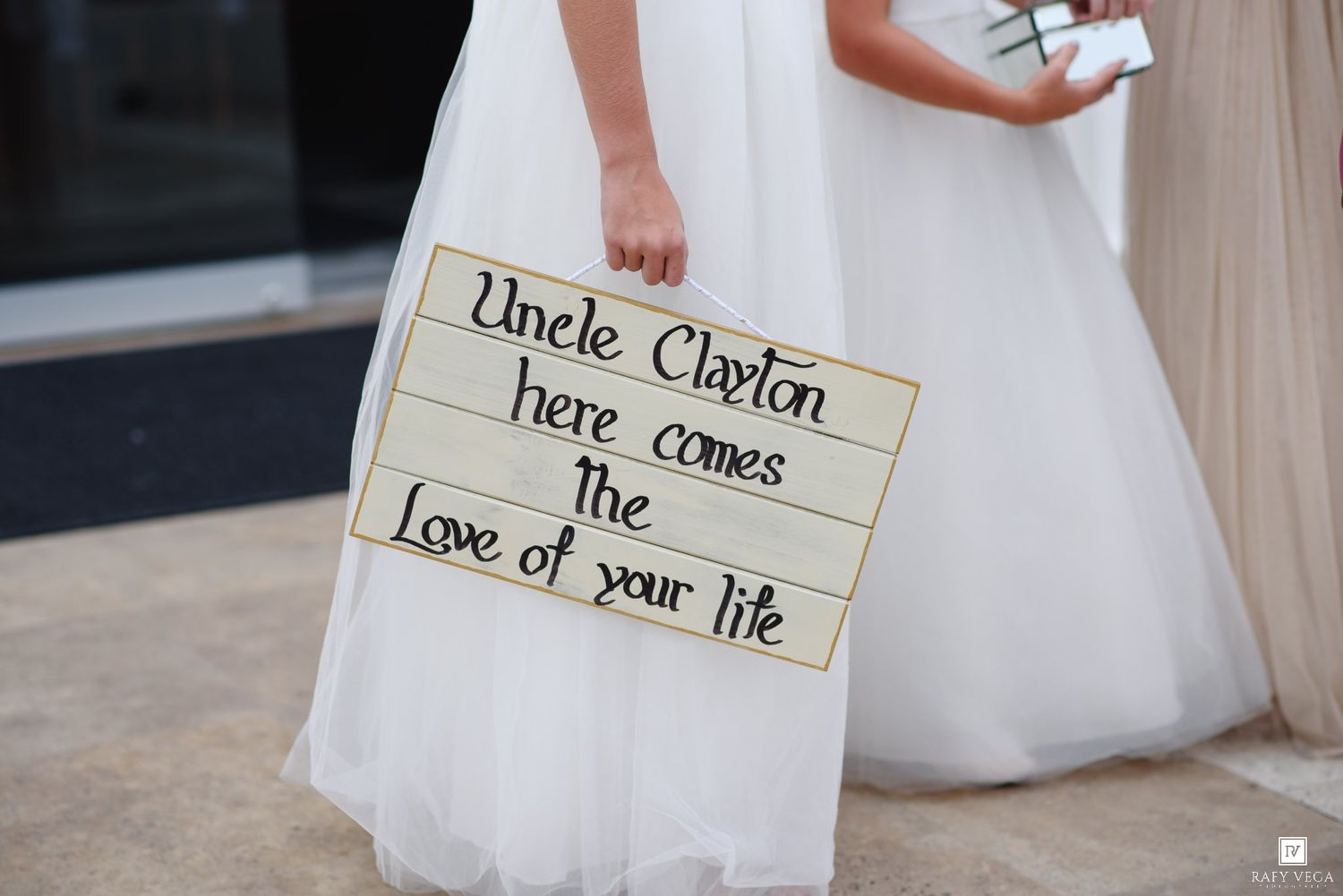 Flower girl holding a sign reading Uncle Clayton here comes the love of your life.