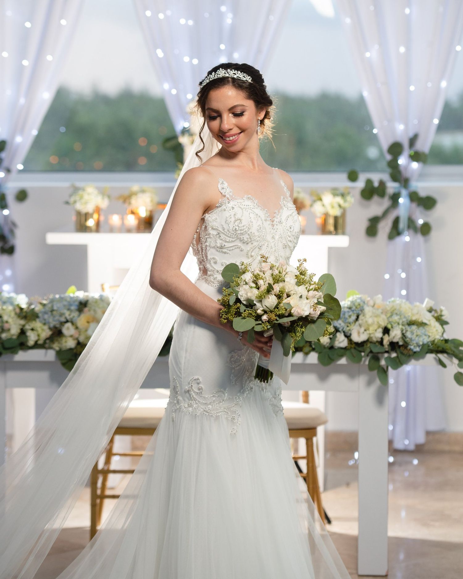 Bride in lace mermaid wedding gown with veil and tiara holds white bouquet in elegant decorated ceremony venue.