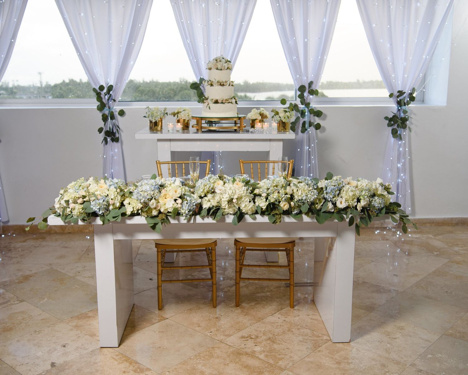 Elegant wedding sweetheart table with white floral garland, gold chairs, sheer drapes, and tiered cake display in background.