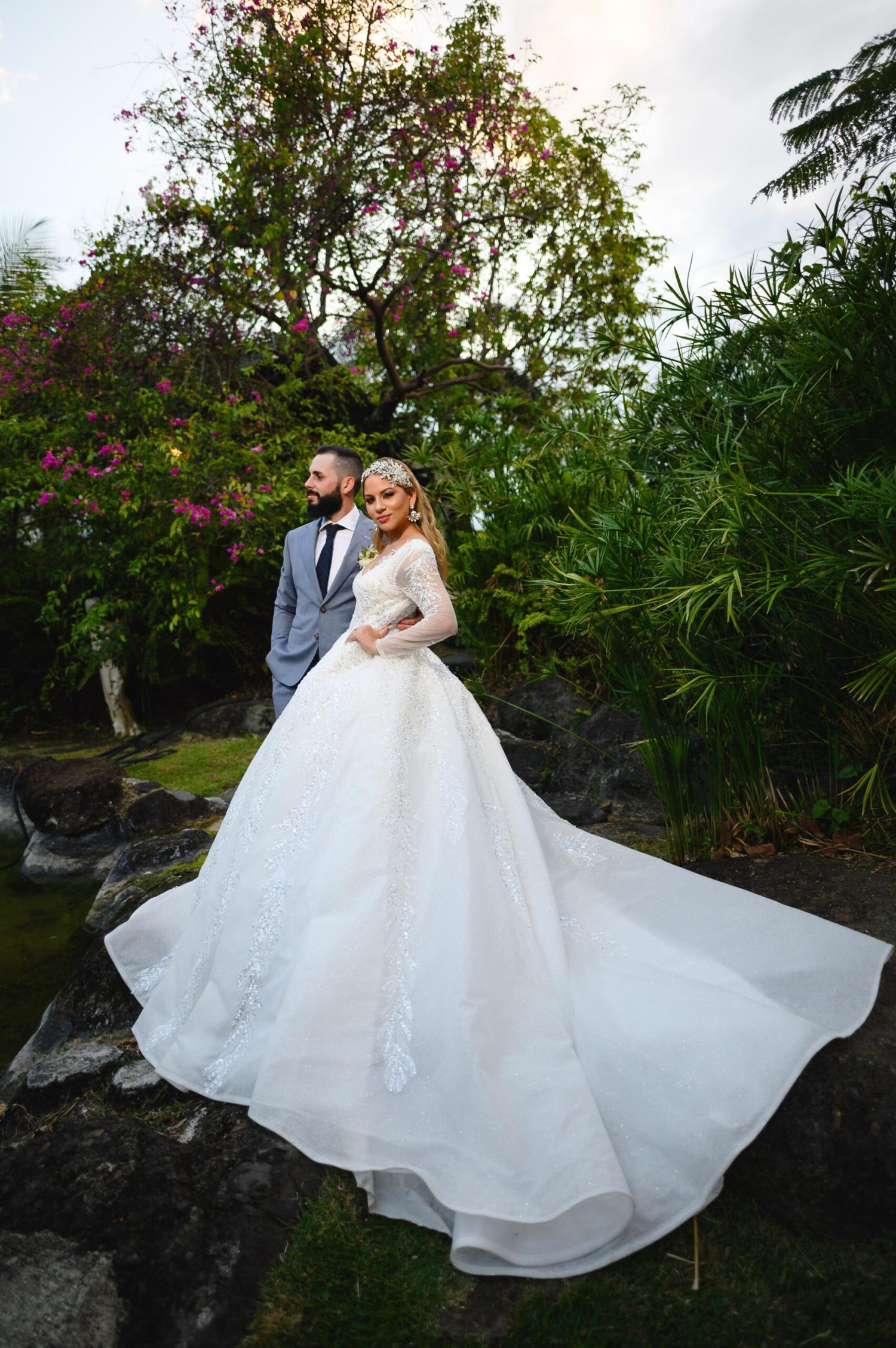 Bride in long-sleeve lace ball gown with flowing train stands with groom in gray suit in lush garden.