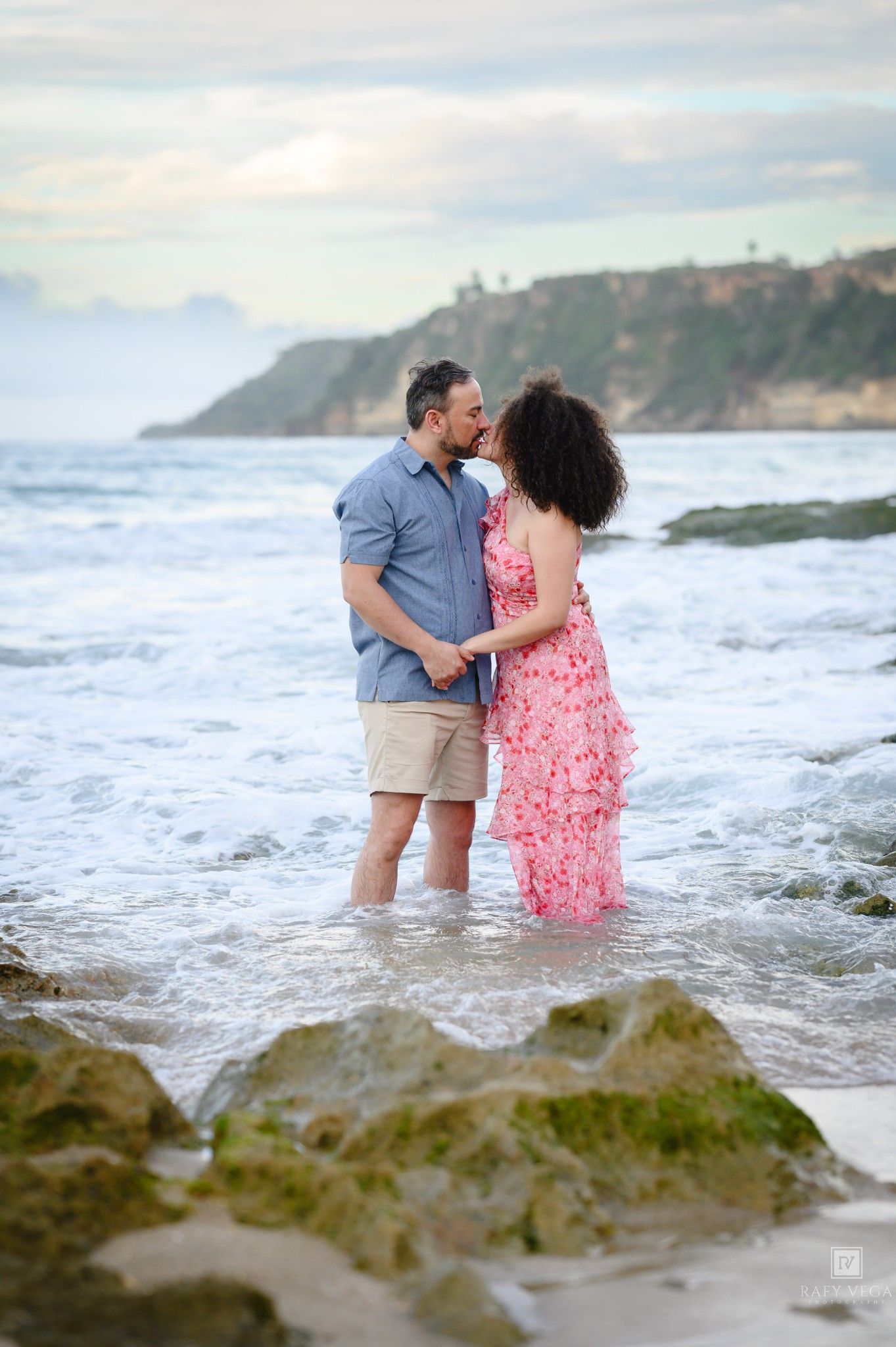 Couple sharing a romantic kiss while standing in ocean waves on a rocky beach with a scenic coastal cliff in the background.