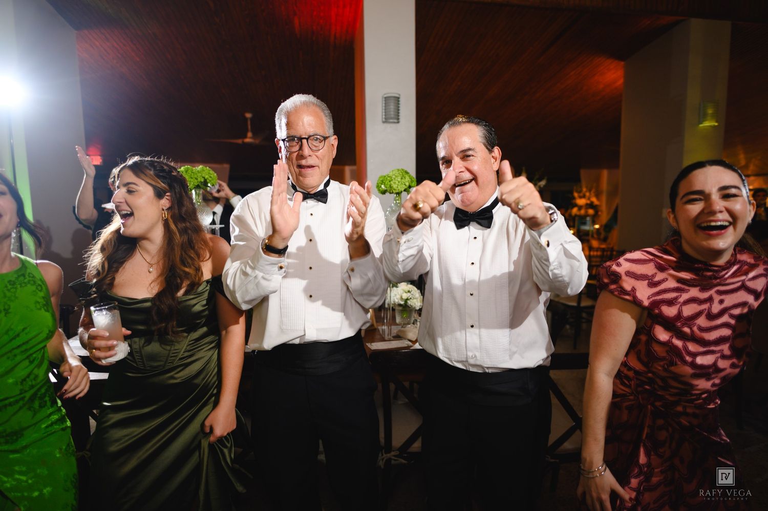 Four guests in formal attire laugh and clap joyfully at an elegant wedding reception celebration.