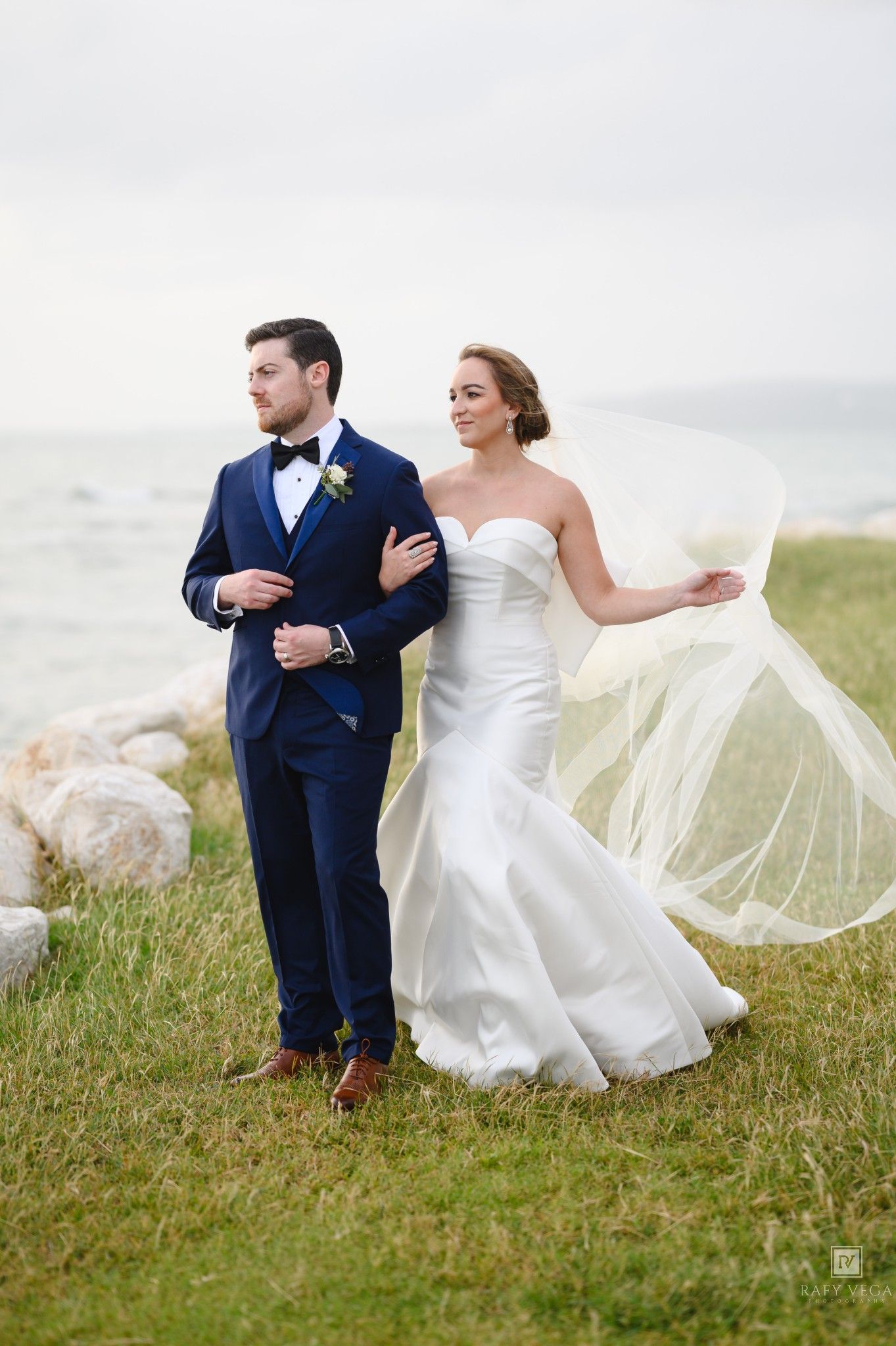 Bride in white strapless gown with flowing veil and groom in navy blue tuxedo pose near rocky shoreline.