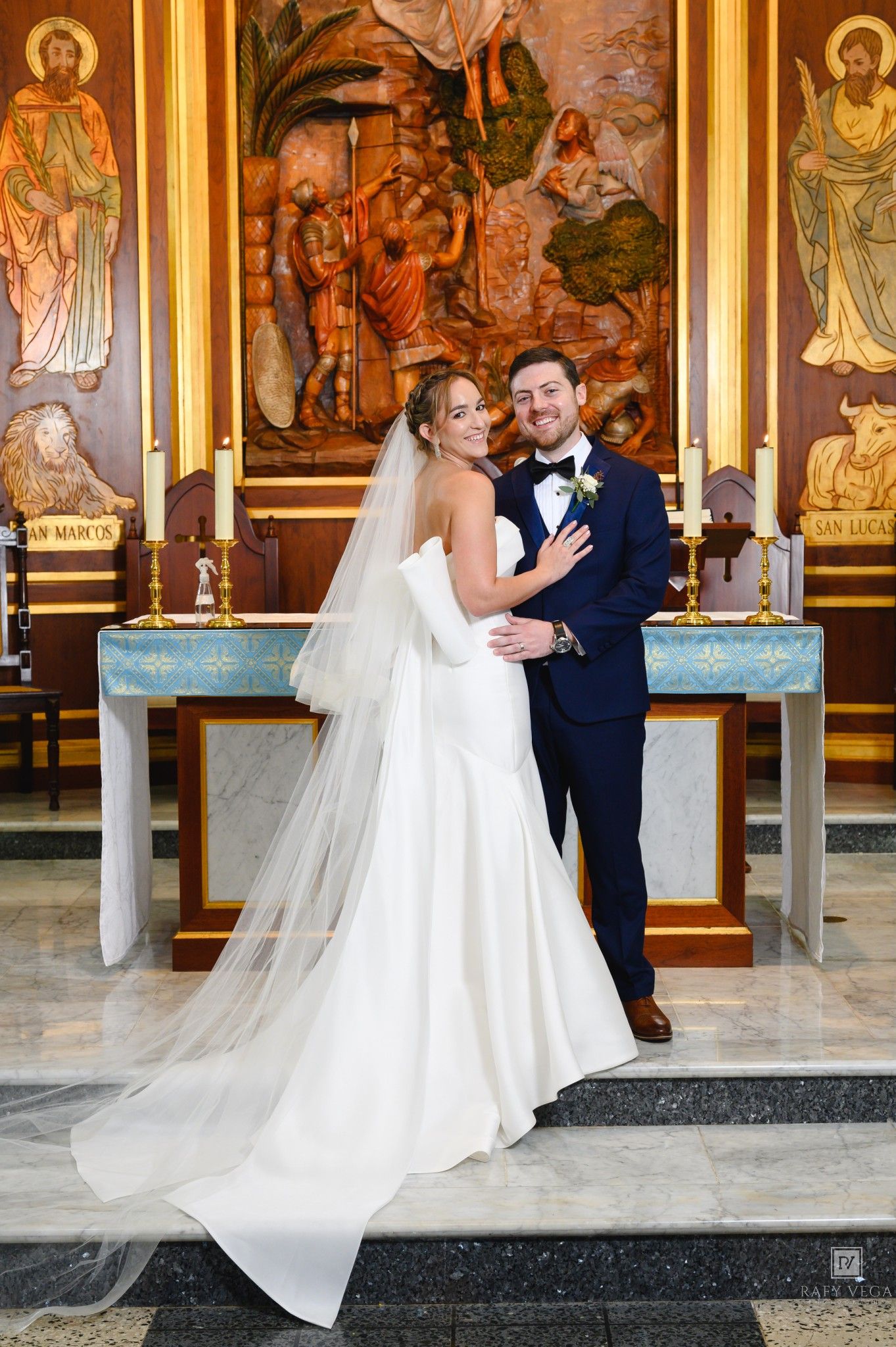 Bride in strapless white gown with veil embraces groom in navy suit at church altar after wedding ceremony.