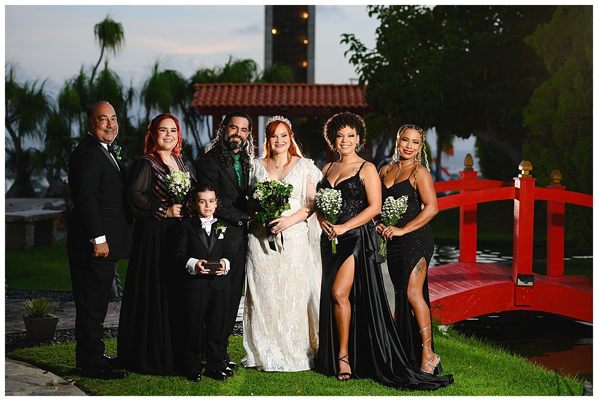 Wedding party poses outdoors near red bridge, bride in white gown with bridesmaids in black dresses holding bouquets.