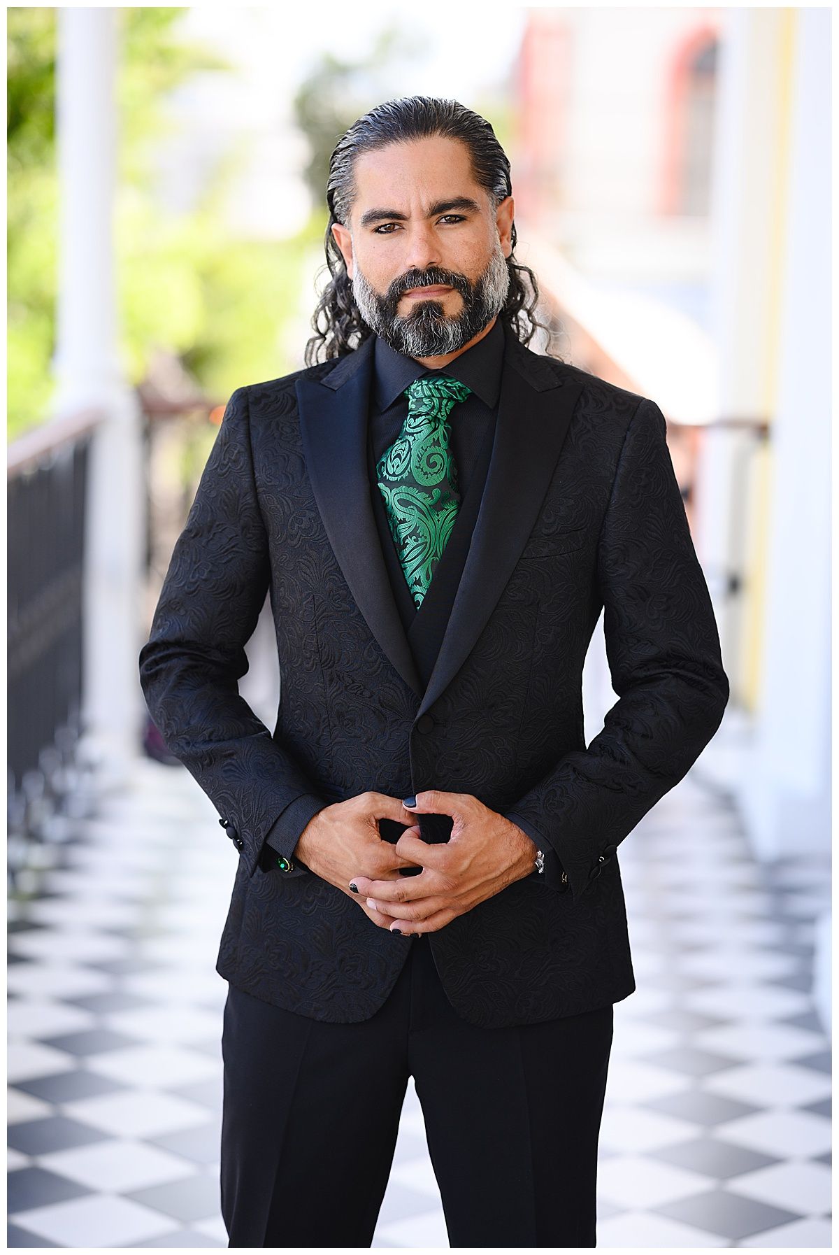 Man in black floral tuxedo jacket with green paisley tie, wearing formal attire outdoors on checkered floor.